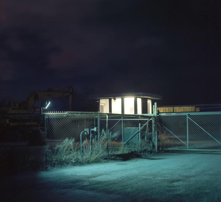 A small illuminated building stands behind a chain-link fence at night, with a large yellow excavator visible in the background.