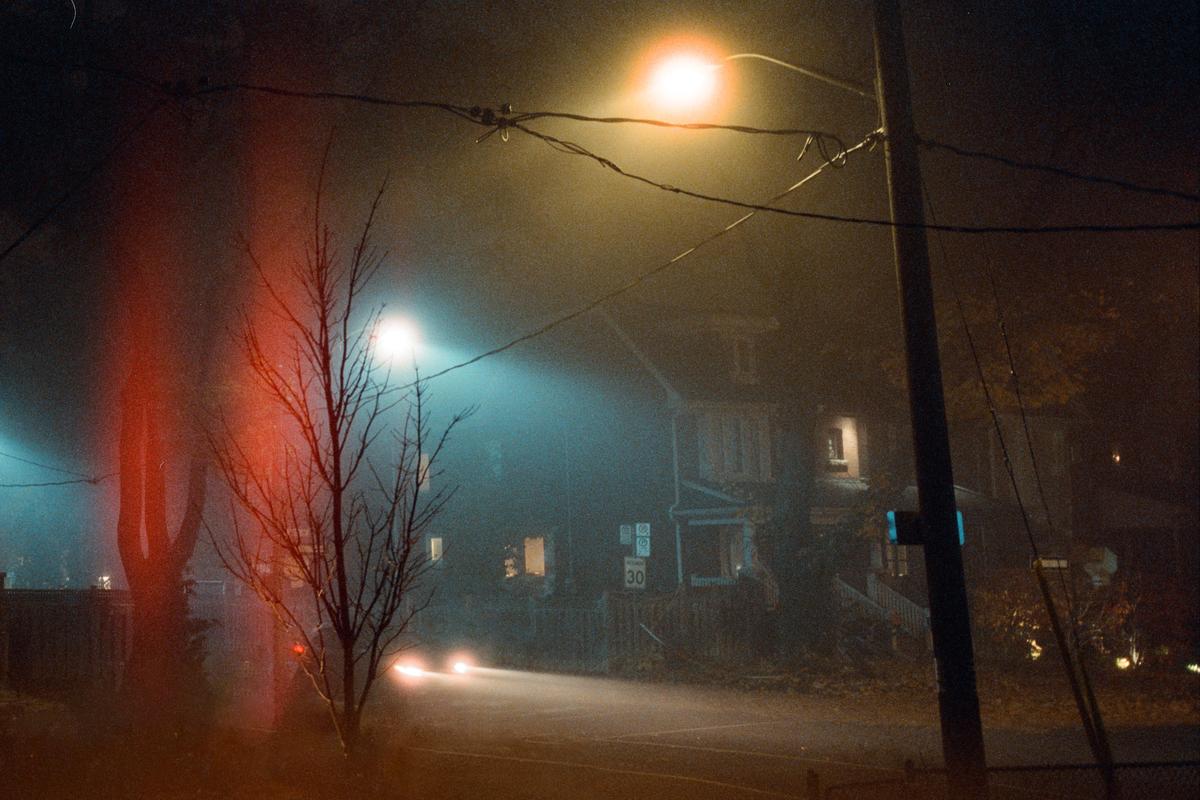 A street at night is illuminated by streetlights and car headlights, with a house visible through the fog.