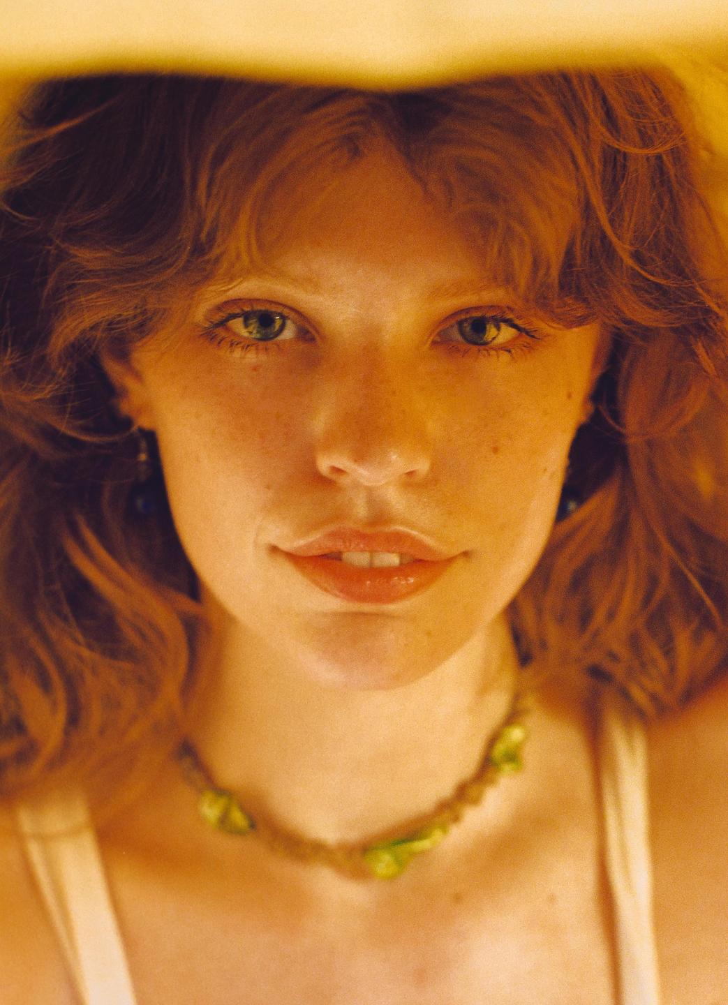 A woman with red hair and a green necklace looks directly at the viewer.