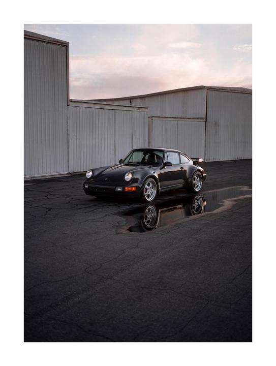 A gray car is parked on asphalt next to white buildings, with its reflection visible in a puddle.