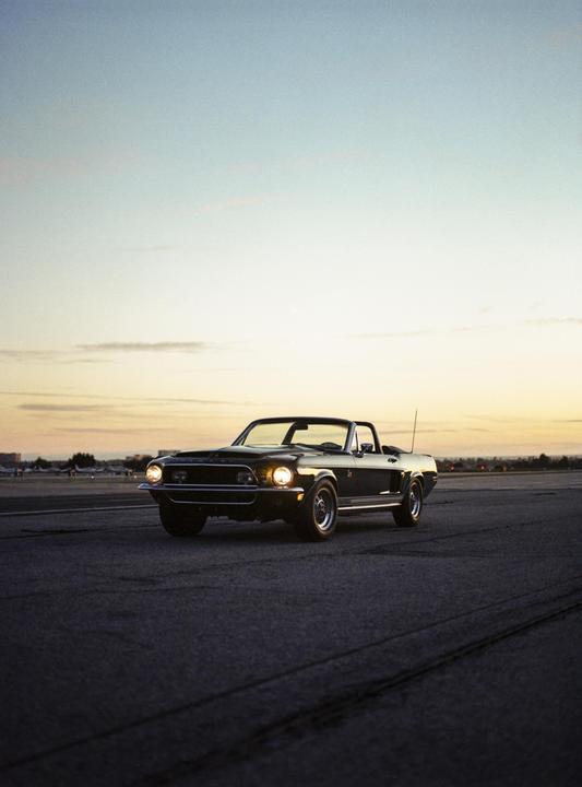 A black convertible Mustang is parked on an airfield at dusk.