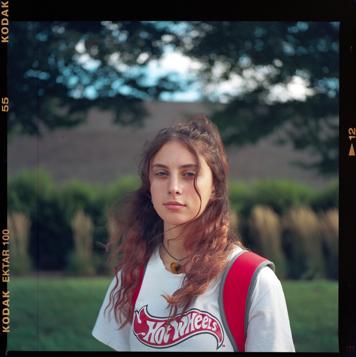A young woman with long, curly hair and a red backpack stands outdoors, wearing a white t-shirt with a Hot Wheels logo.