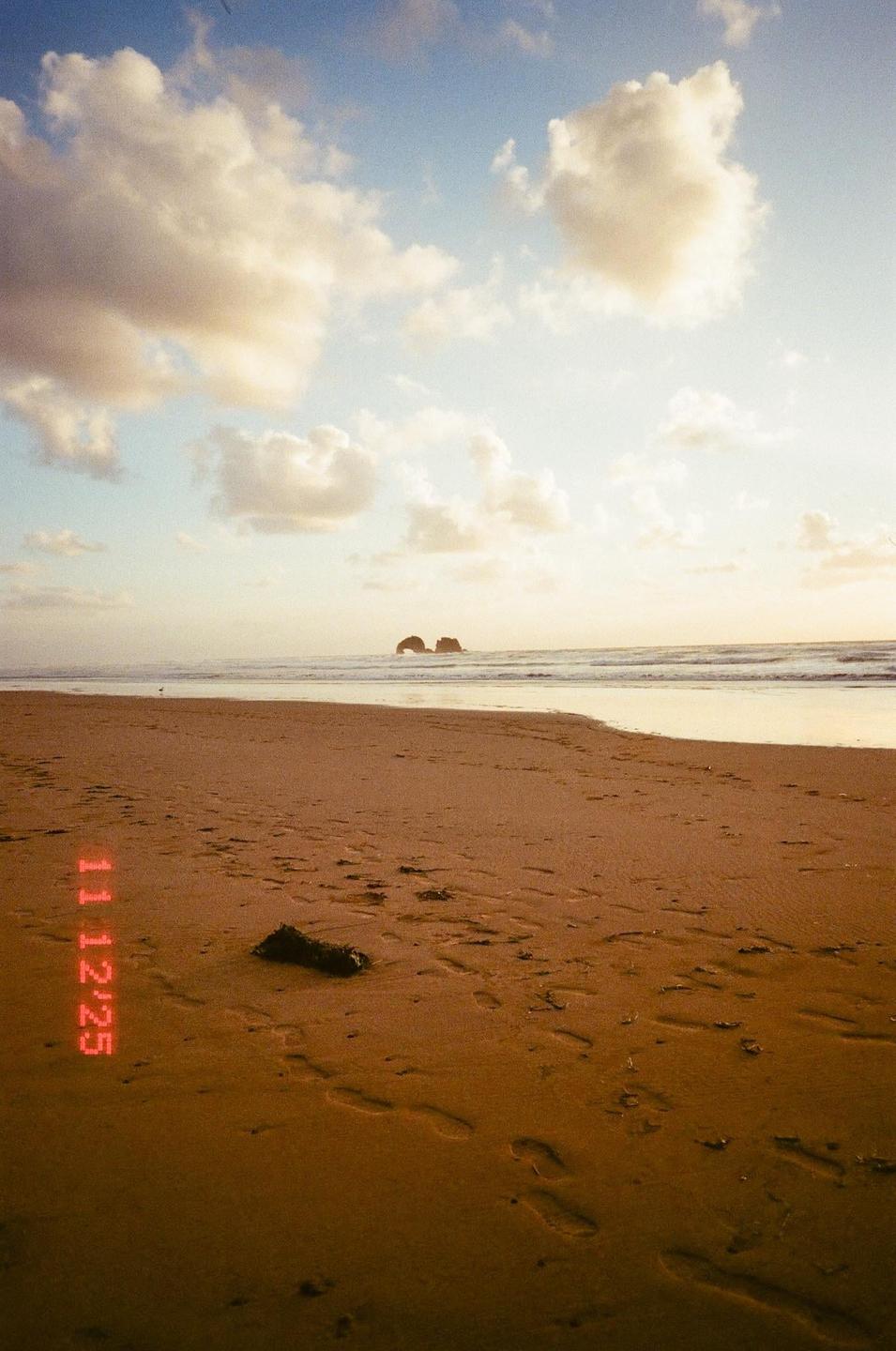 A sandy beach with footprints leads to the ocean where two rock formations are visible under a cloudy sky.