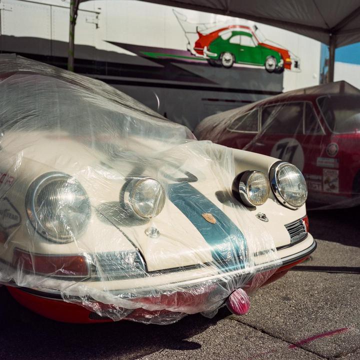 Two vintage cars are covered with plastic sheets, with a painted car graphic on a white wall in the background.