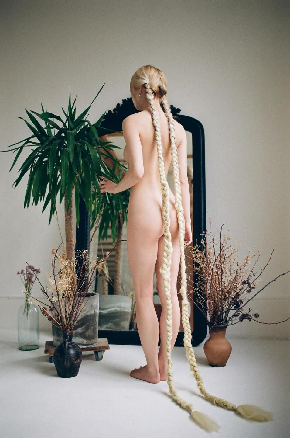A nude woman with long braided hair stands with her back to the camera, looking into a mirror next to a large plant.