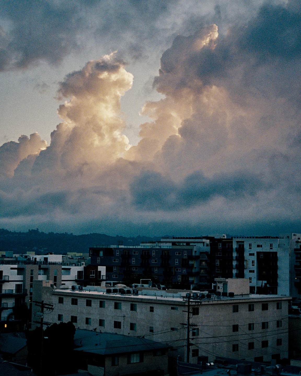 A dramatic sky with large, illuminated clouds hangs over a city with apartment buildings and distant hills.