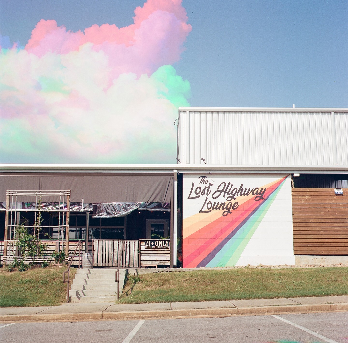 A building with a rainbow mural and a sign for "The Lost Highway Lounge" stands under a sky with colorful clouds.