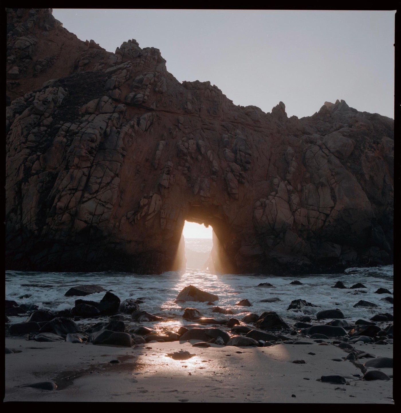 Sunlight streams through a natural arch in a rocky cliff onto a sandy beach with waves.