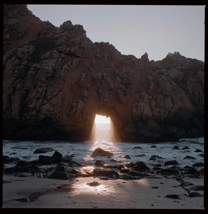 Sunlight streams through a natural arch in a rocky cliff onto a sandy beach with waves.