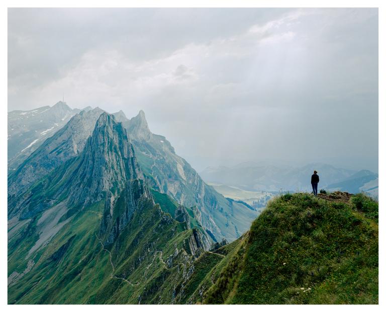 A person stands on a grassy mountain peak overlooking a valley and other mountains under a cloudy sky.