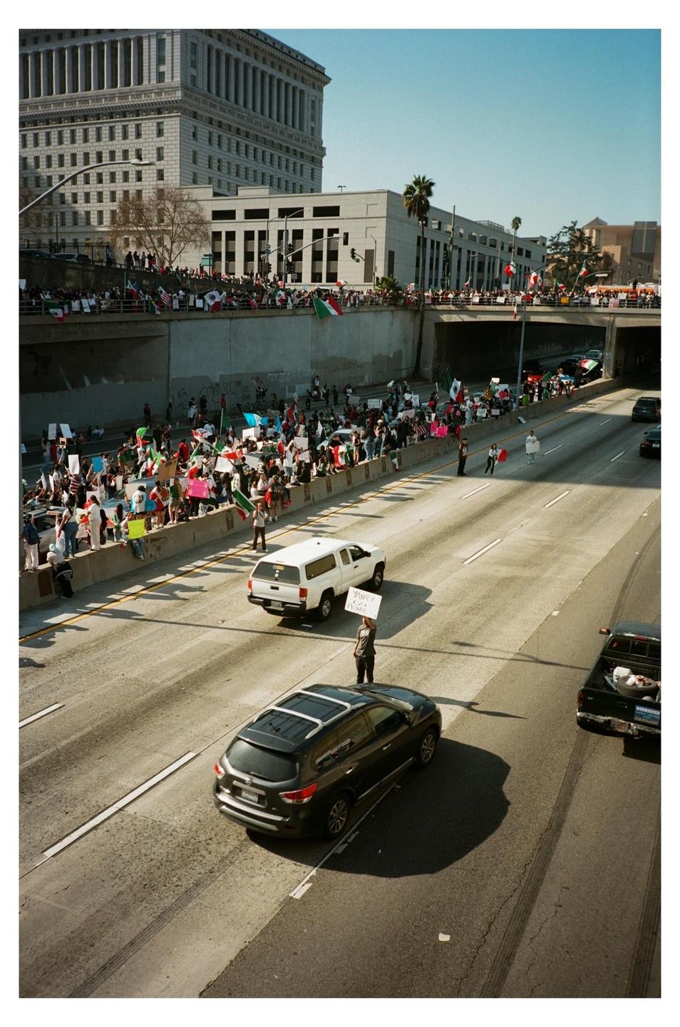 A large crowd of people with flags and signs are gathered on a bridge overlooking a highway with cars and a person holding a sign.