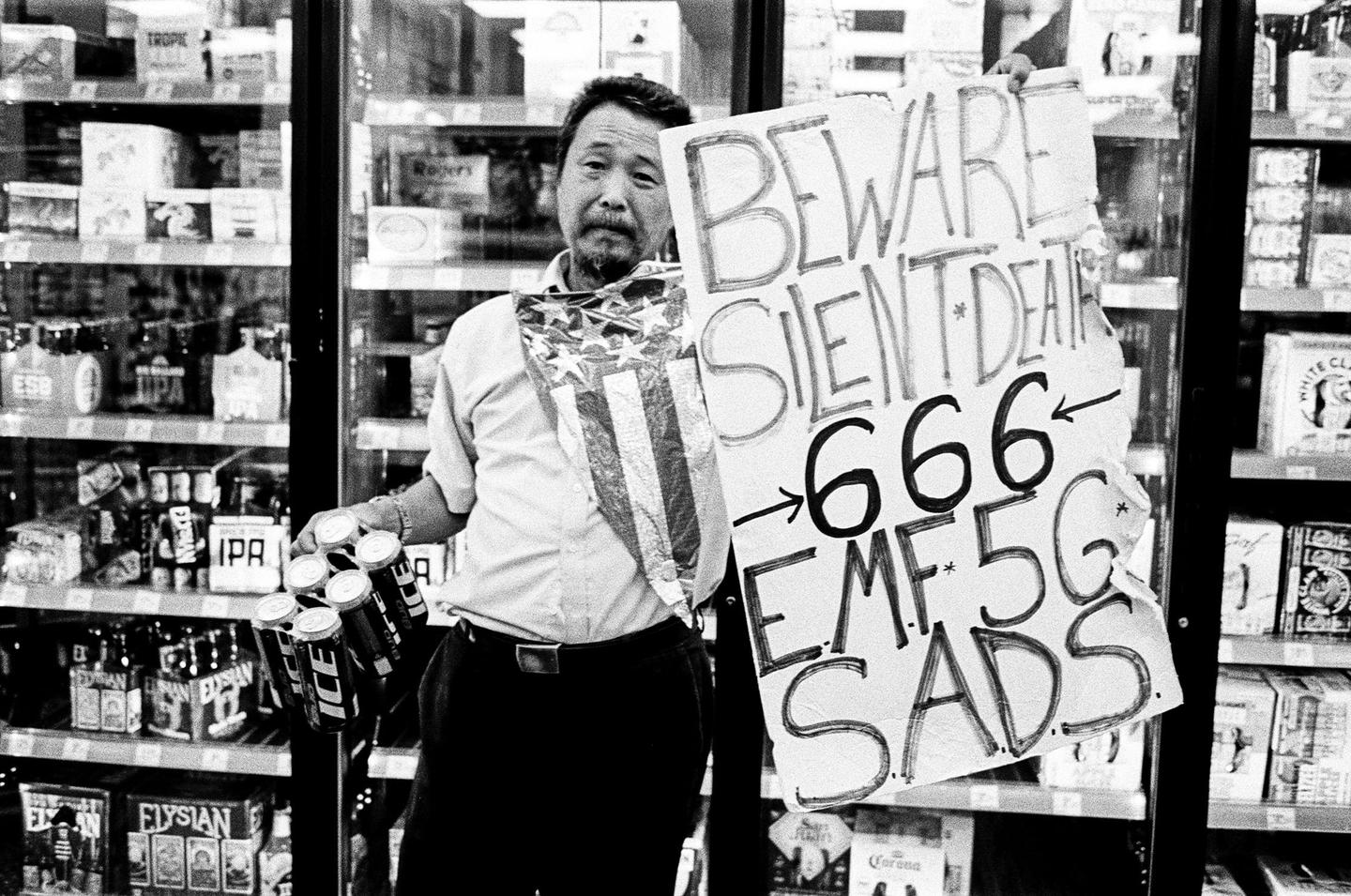 A man holds a sign and a six-pack of beer in front of a refrigerated display case in a store.