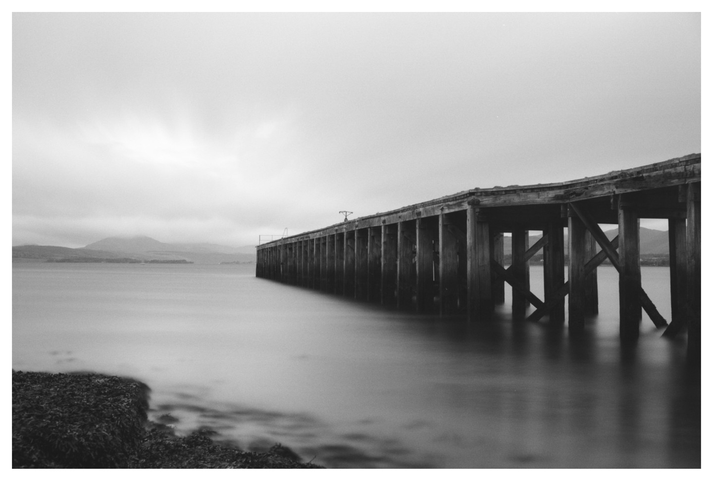 A long exposure captures a wooden pier extending into calm water under a cloudy sky with distant mountains.