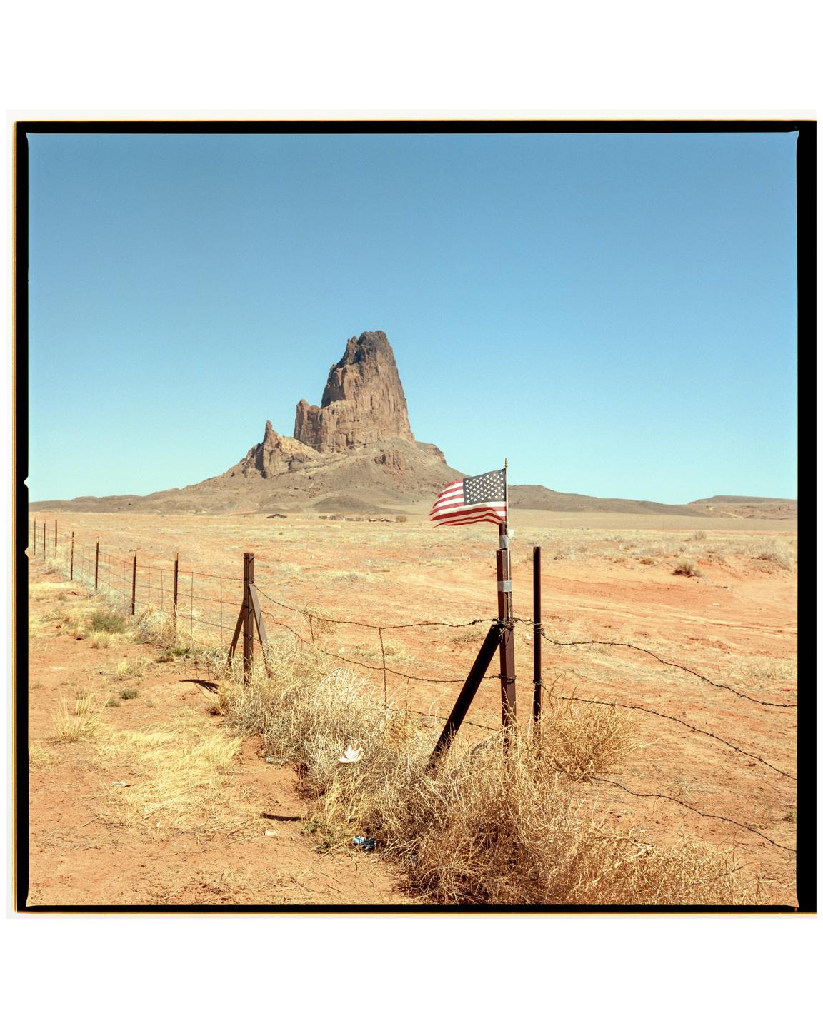 An American flag is attached to a fence post in a desert landscape with a large rock formation in the background.