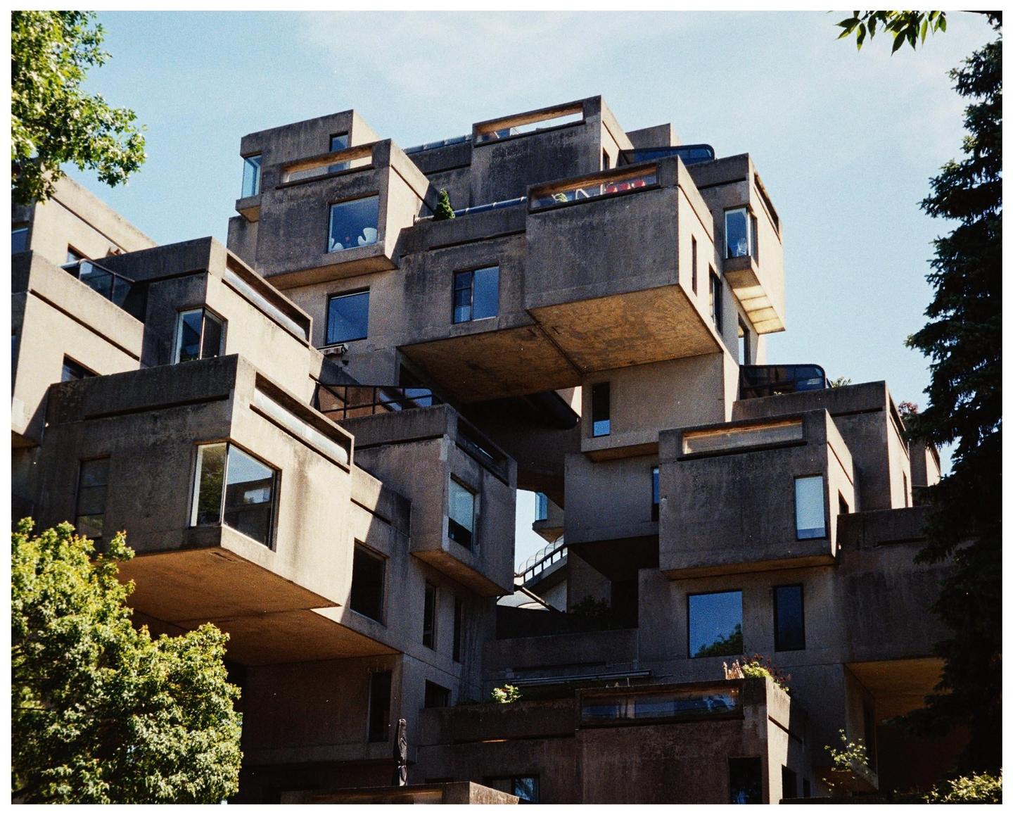 A complex concrete building with many windows and balconies is surrounded by green trees under a clear sky.