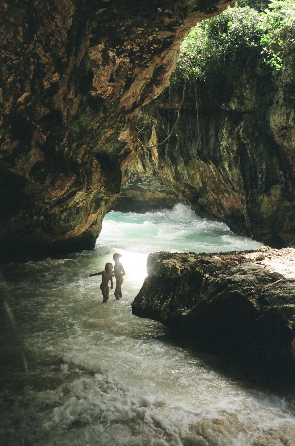 Two children stand in the water inside a sea cave with waves crashing in the background.