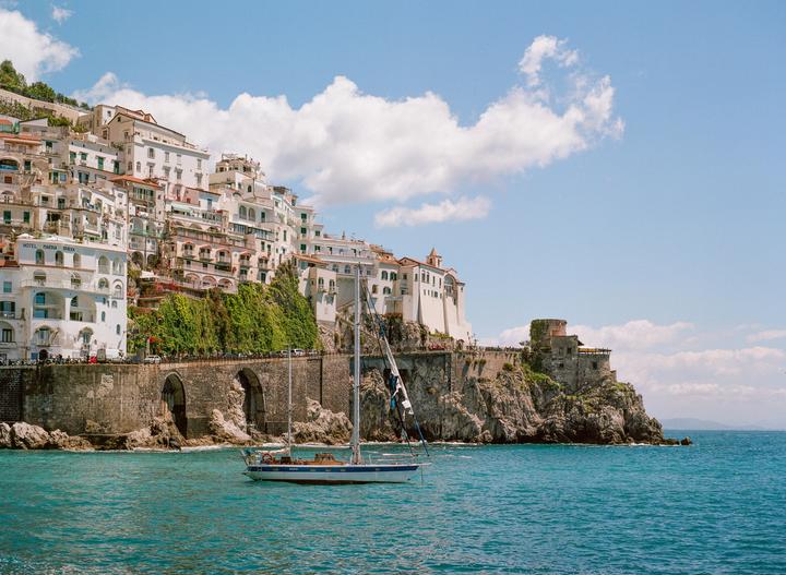 A sailboat floats on the water in front of a town built into a cliffside.