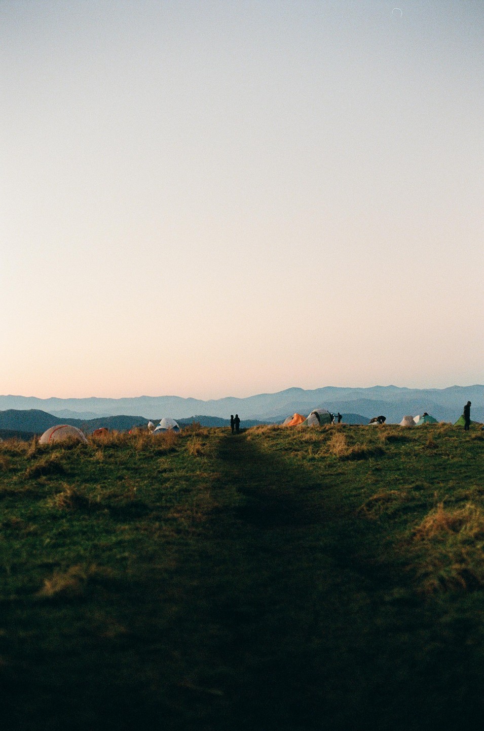 A group of tents is pitched on a grassy mountain ridge with a couple standing on a path in the distance.