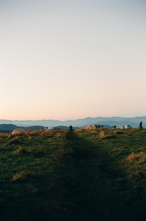 A group of tents is pitched on a grassy mountain ridge with a couple standing on a path in the distance.