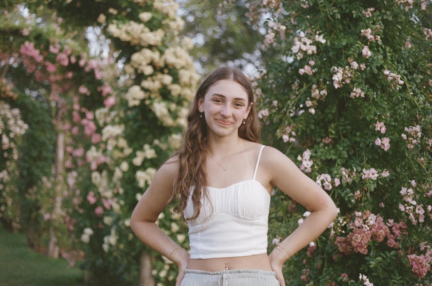 A young woman stands smiling in a garden with flowering bushes behind her.