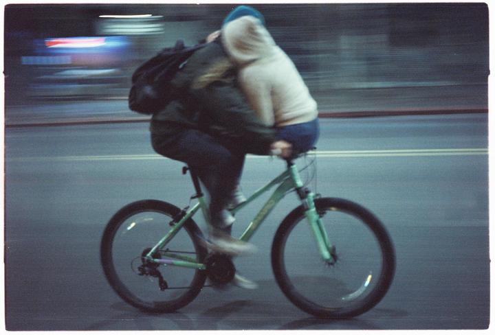 Two people ride a bicycle at night, with one person sitting on the handlebars and hugging the other.