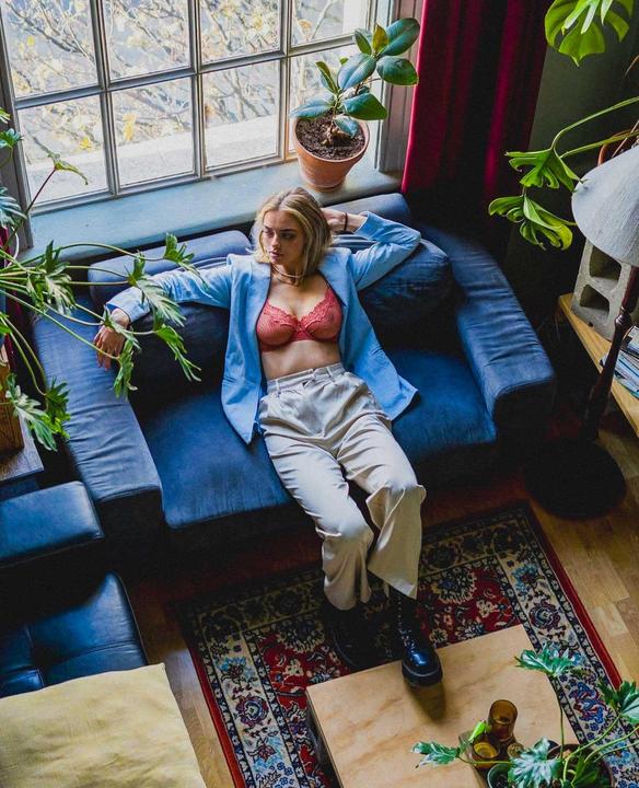 A woman in a blue blazer and red bra is reclining on a sofa in a room with plants and a window.