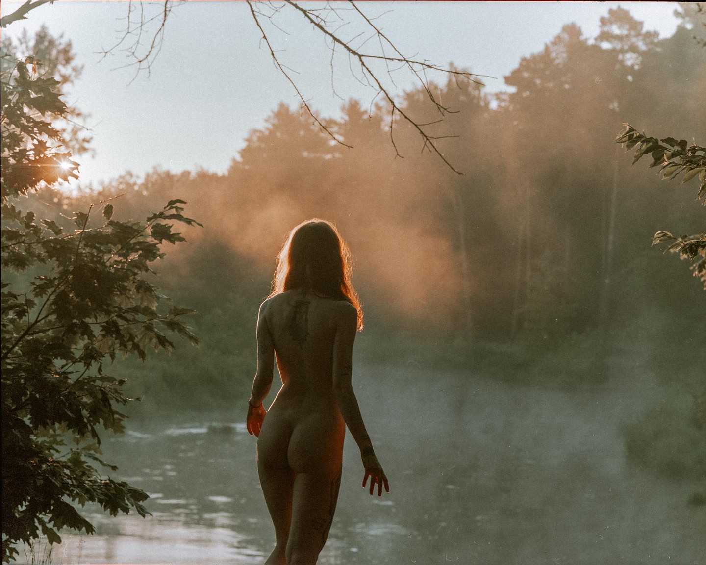 A nude woman stands at the edge of a misty river with a forest in the background.
