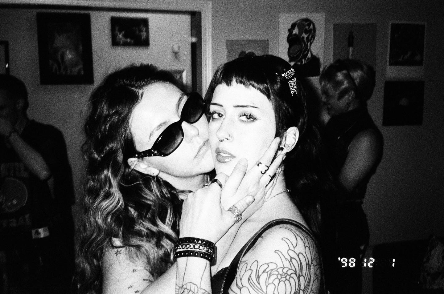 A woman wearing sunglasses kisses another woman on the cheek, who is looking at the camera.
