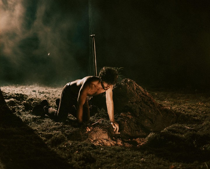 A man kneels on the ground next to a pile of dirt, reaching into a small hole with his hand.