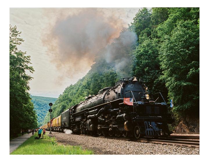 A large black steam locomotive with an American flag on its front travels along a track next to a forested hillside.