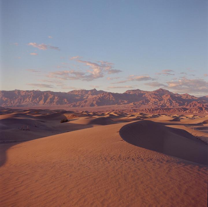A vast desert landscape features sand dunes in the foreground and mountains in the distance under a partly cloudy sky.