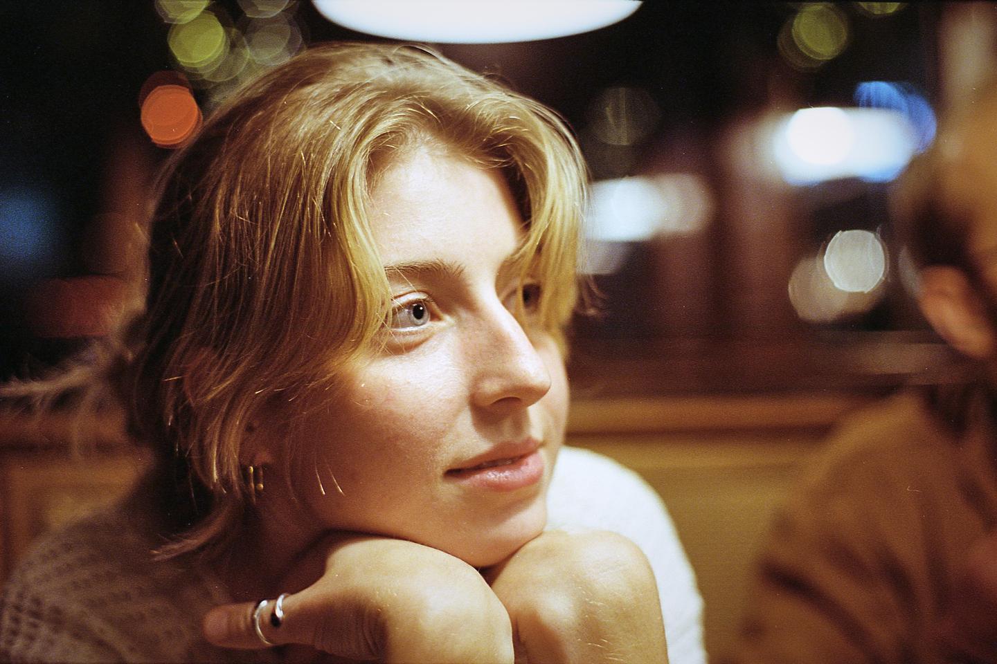A young woman with blonde hair rests her chin on her hands, looking to the right with bokeh lights in the background.