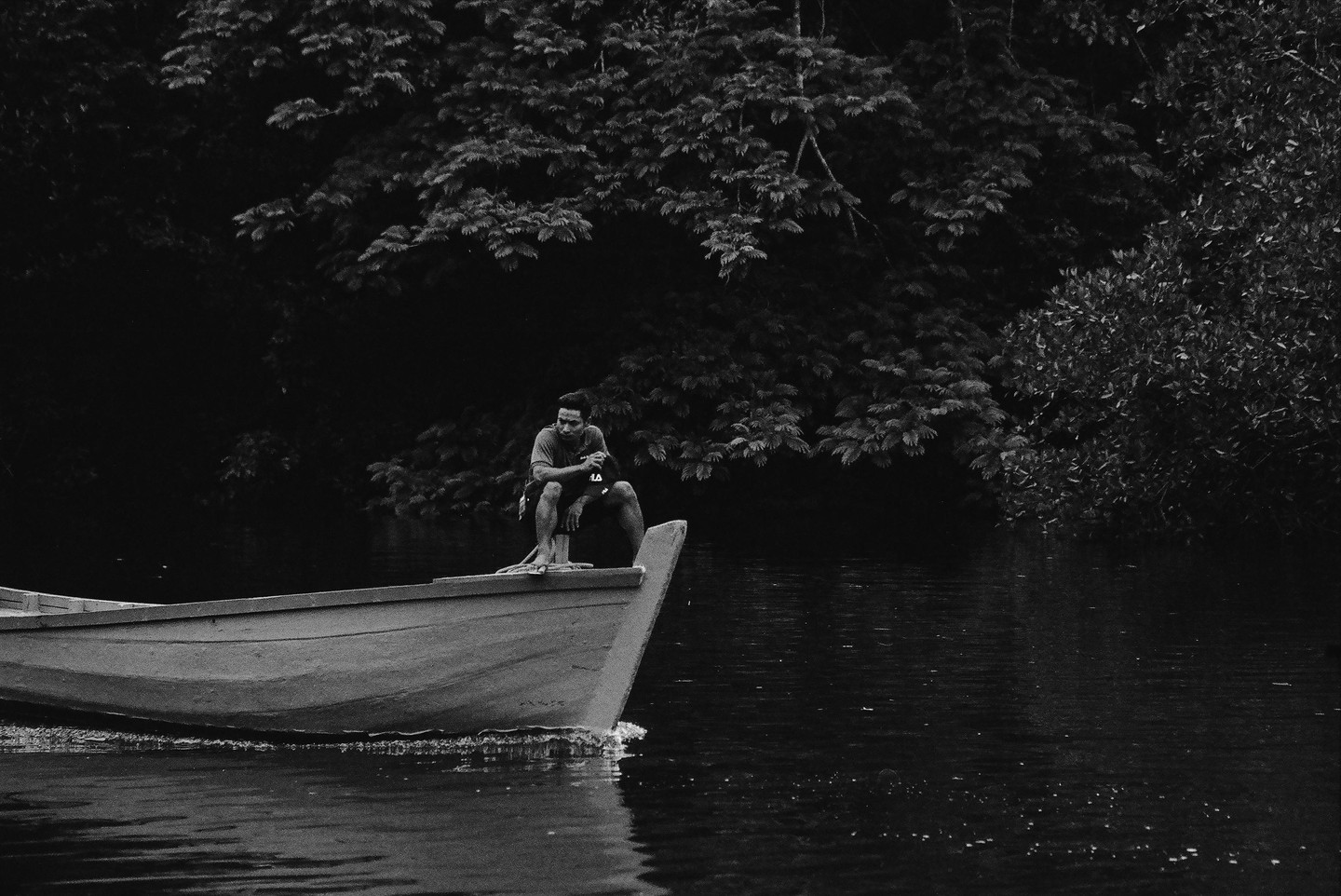 A man crouches at the front of a boat on dark water with dense foliage in the background.