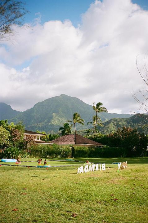 A large green lawn with children playing and a "BIRTHDAY" sign in the foreground, with houses, palm trees, and mountains in the background.