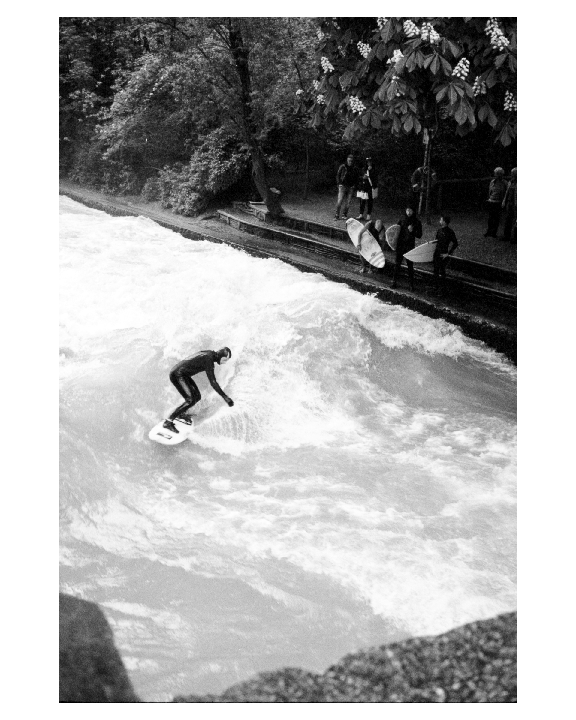 A surfer in a wetsuit rides a wave in a river while people watch from the bank.