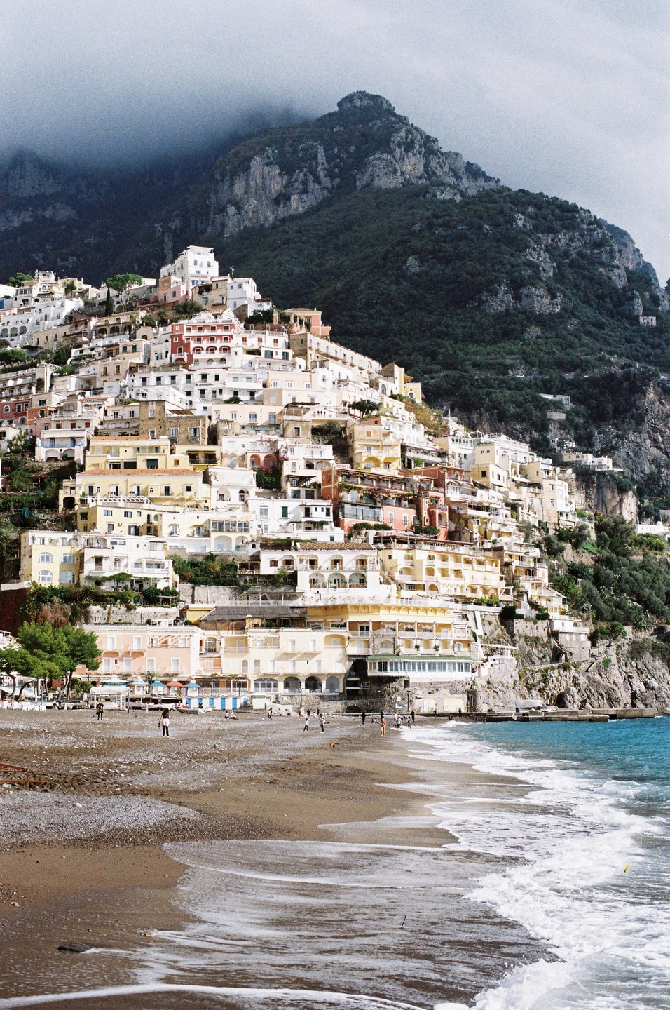 A hillside town with many buildings is visible from a beach with waves crashing on the shore.