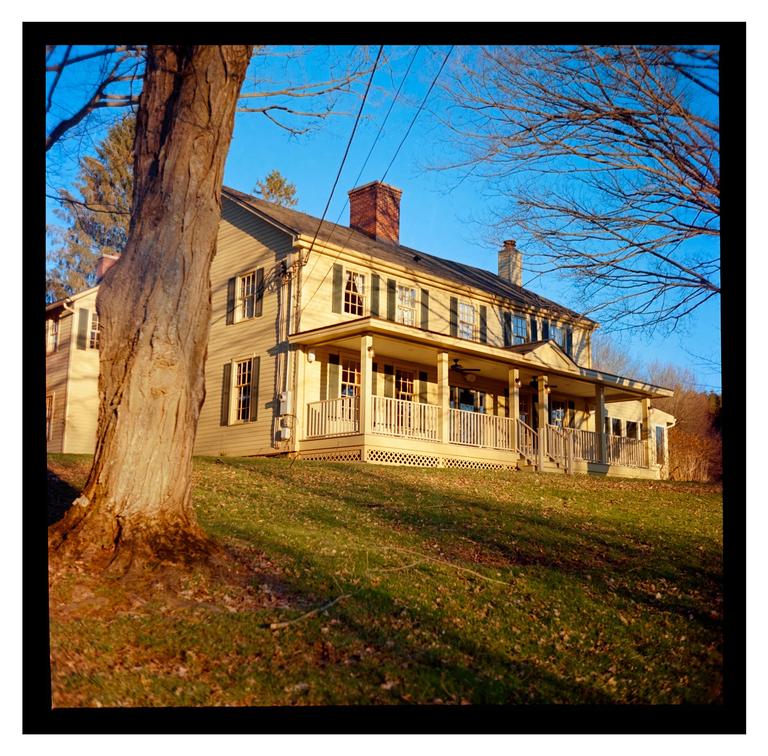 A large house with a long porch and green shutters sits on a grassy hill under a clear blue sky.