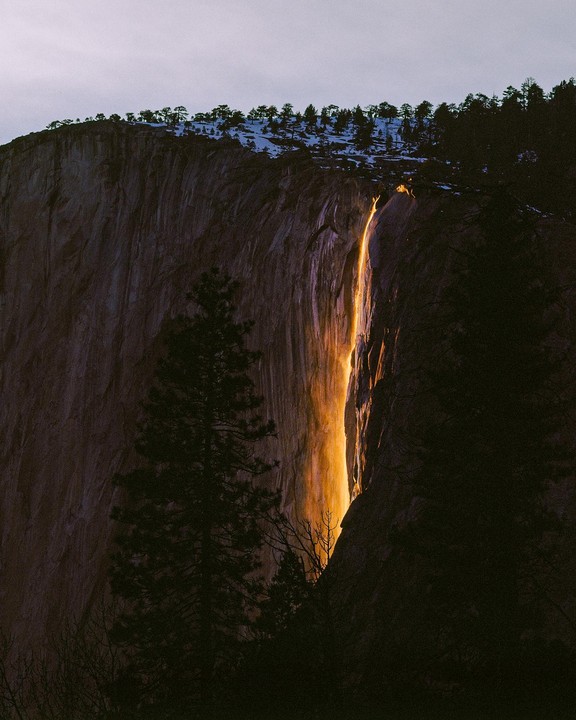 A waterfall glows orange as it cascades down a dark cliff face, with snow-dusted trees visible at the top.