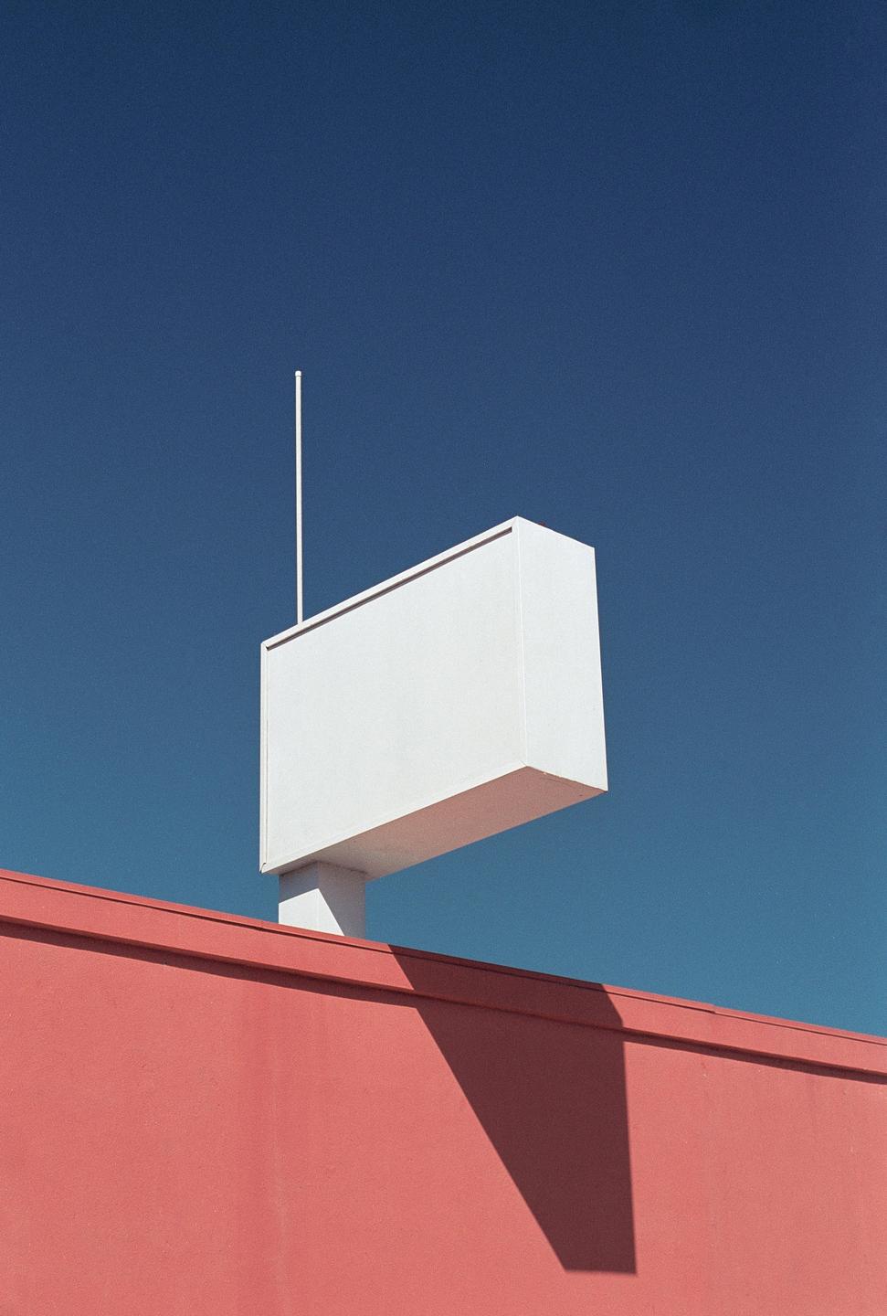 A white rectangular sign with a pole on top is mounted above a red building against a clear blue sky.