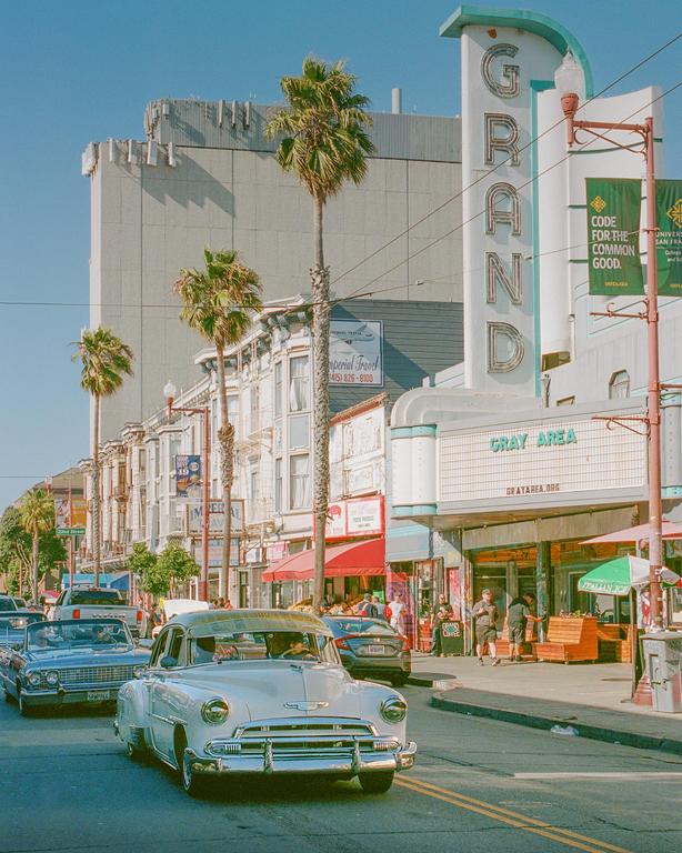 A vintage car drives down a street lined with palm trees and buildings, including the Grand Theater.