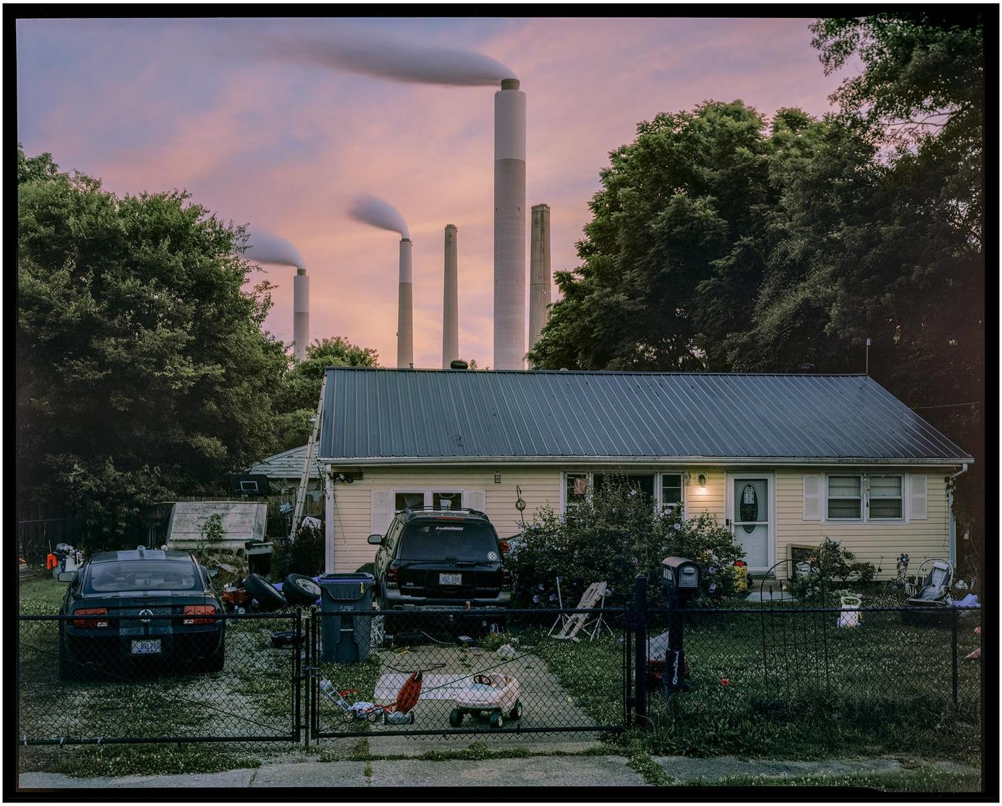 A small house with two cars in the driveway is visible behind a fence, with large industrial smokestacks emitting smoke in the background.
