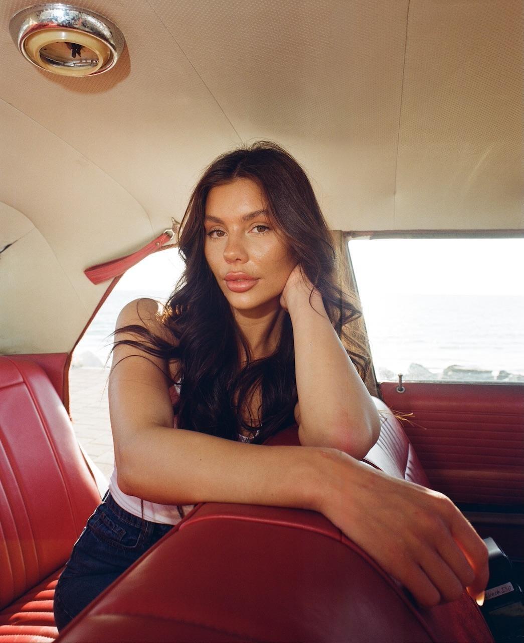 A woman with long, dark hair sits in the red interior of a car, looking towards the viewer.