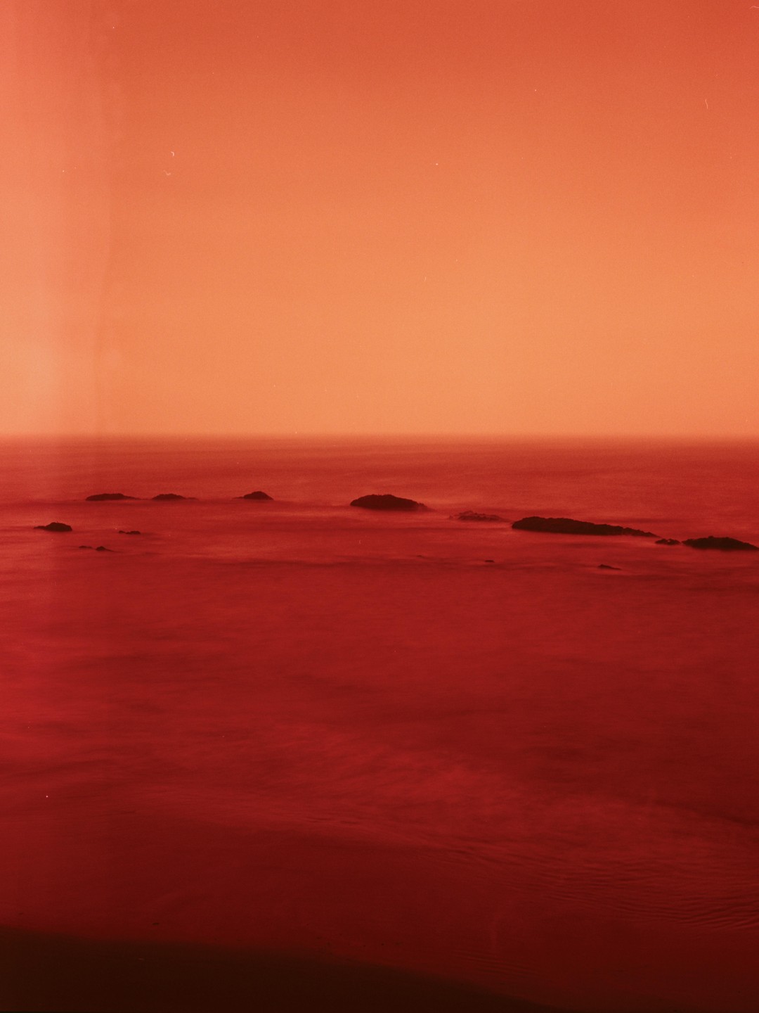 A long exposure photograph of the ocean with several rocks breaking the surface, under an orange sky.
