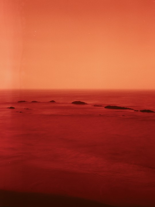 A long exposure photograph of the ocean with several rocks breaking the surface, under an orange sky.