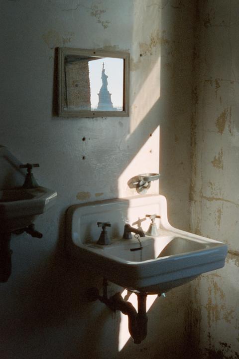 Sunlight illuminates two sinks in a dilapidated room, with the Statue of Liberty visible in a small mirror on the wall.