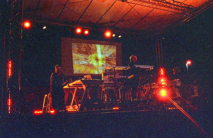 A band performs on a stage at night, illuminated by red lights and a projected image.