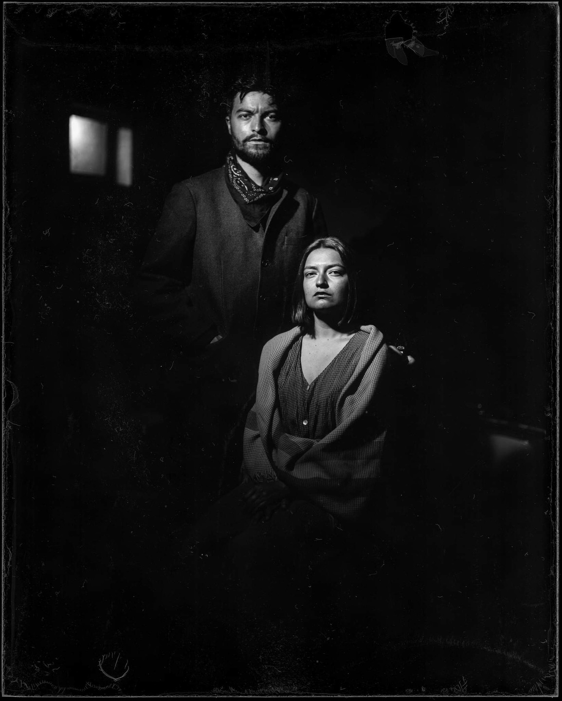 A man stands behind a seated woman, both looking directly at the viewer in a dimly lit room.
