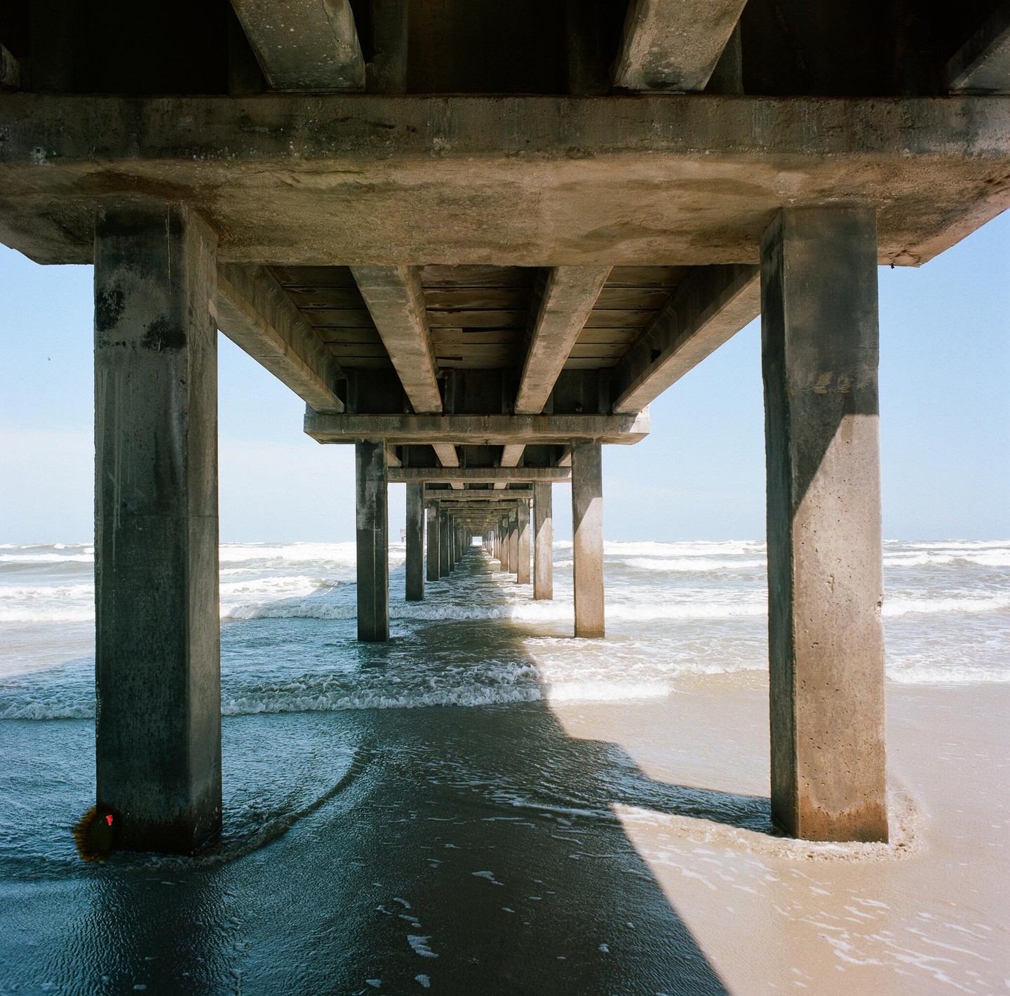 The underside of a concrete pier extends into the ocean, with waves washing around its supports.