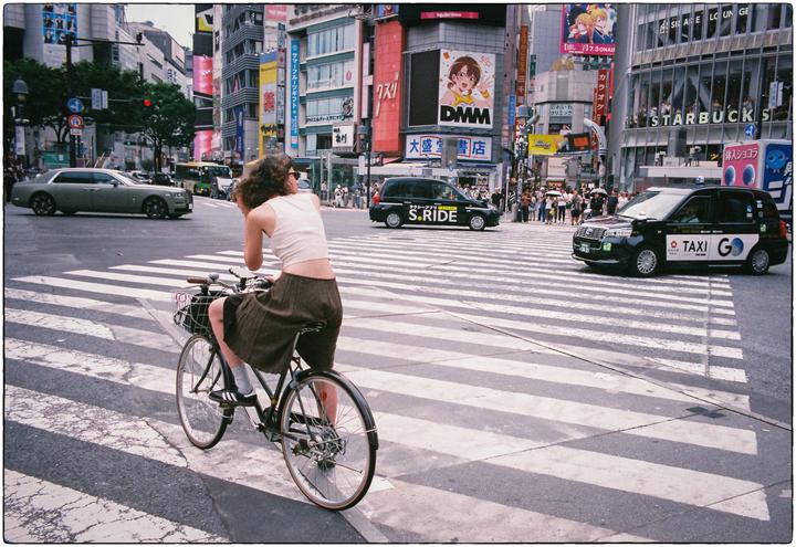 A woman on a bicycle waits at a crosswalk in a busy city intersection with tall buildings and many vehicles.