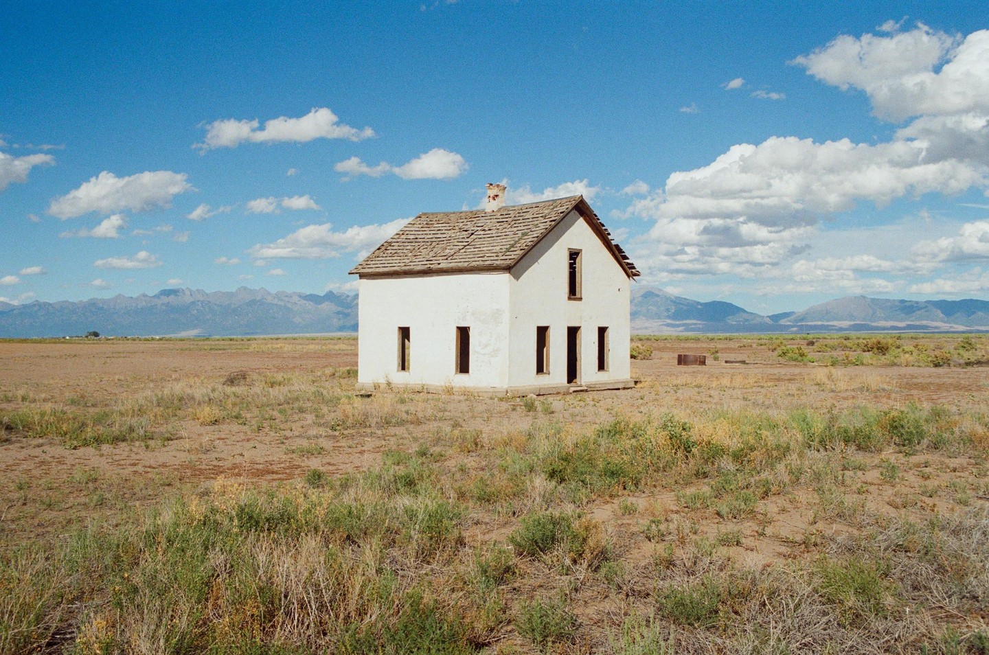 An abandoned white house stands in a dry, grassy field under a blue sky with scattered clouds, with mountains in the distance.